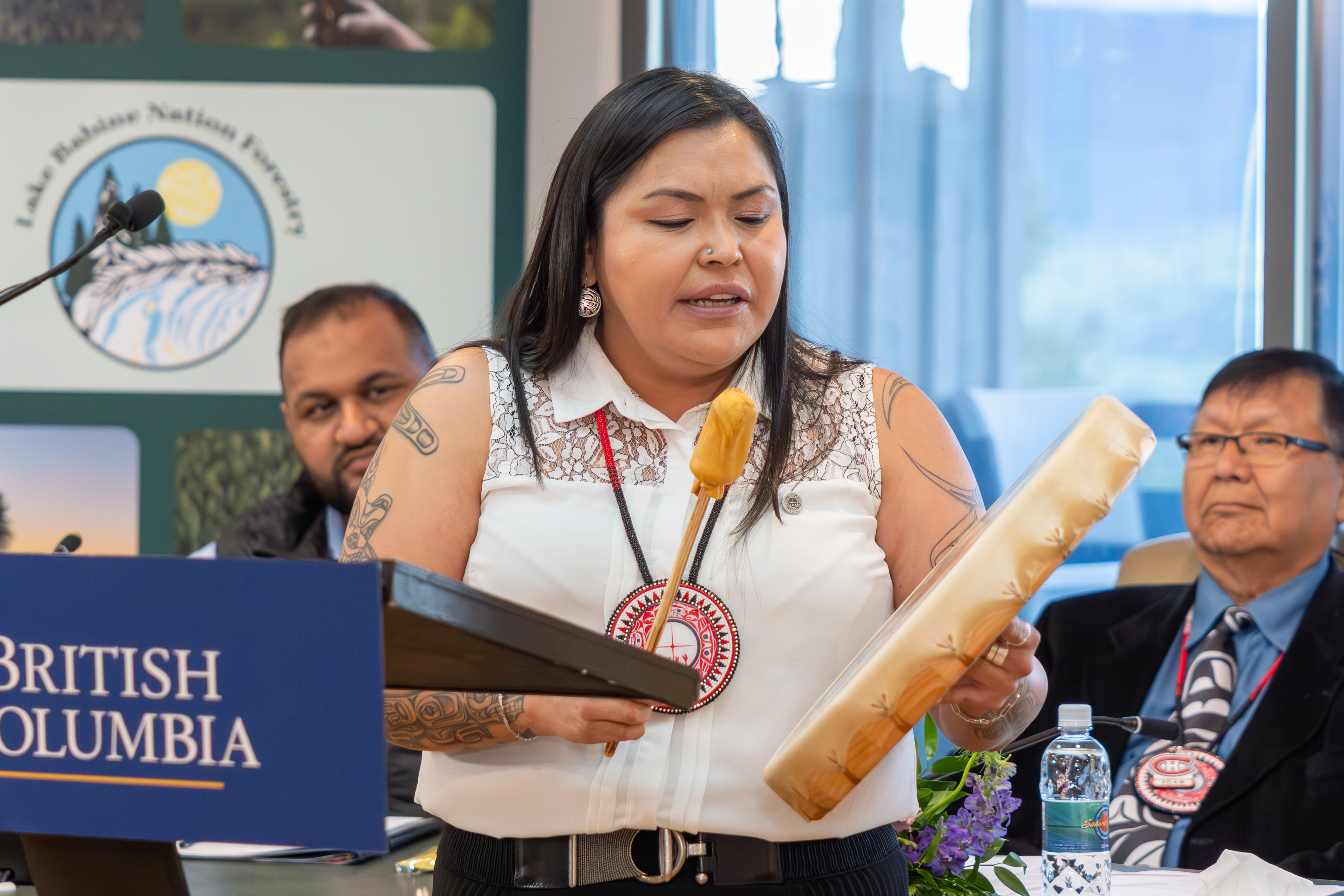 An Indigenous woman holds a drum and drumstick while speaking at a podium with a British Columbia sign; people are seated in the background.