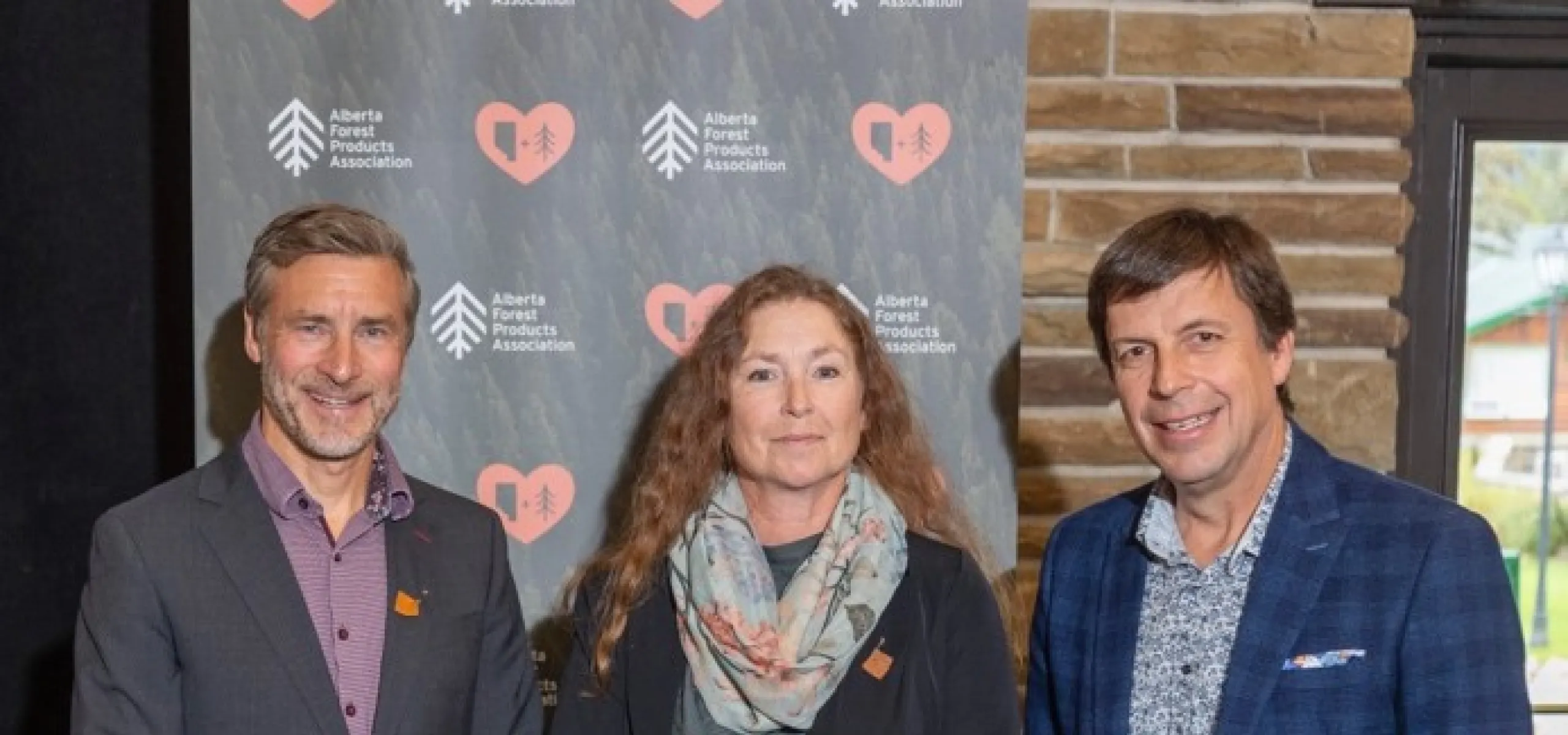 Three individuals pose together, with the middle person holding a red award plaque. They stand in front of a backdrop with logos from the Alberta Forest Products Association.