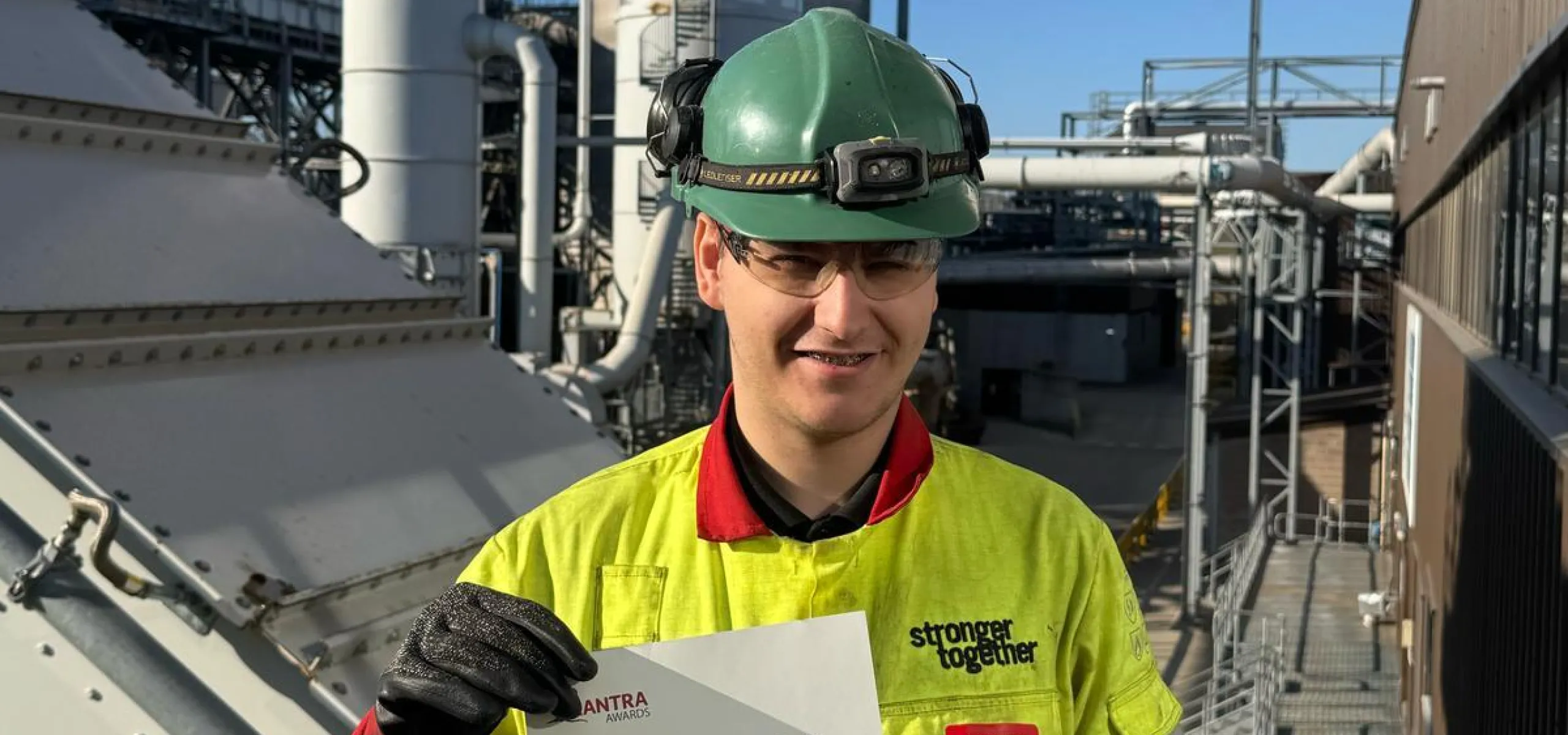A worker wearing safety gear and holding a certificate stands in front of industrial equipment with smoke rising from a stack.